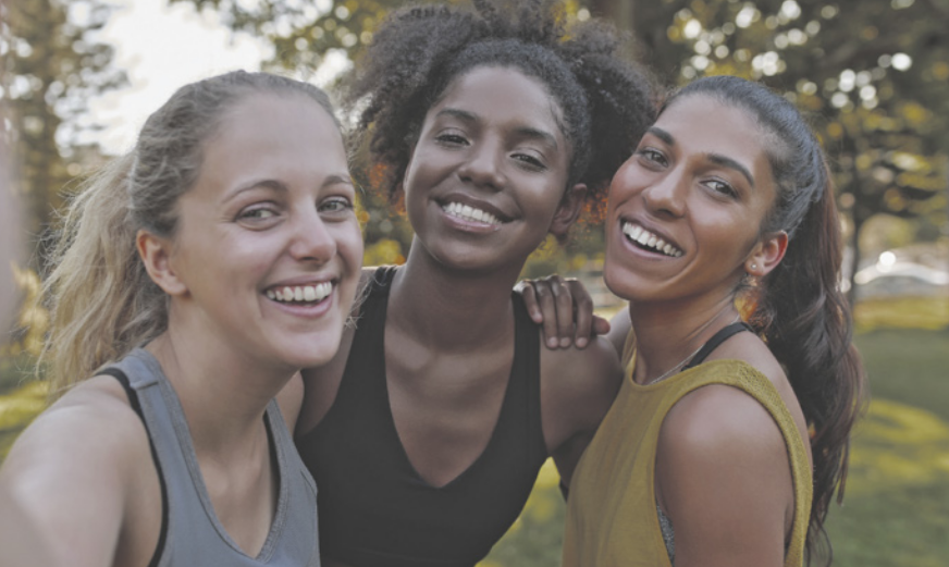 Fotografia de três jovens, uma ao lado da outra, posando para uma foto. Elas estão em um parque, usando roupas de ginástica e sorriem para a foto.