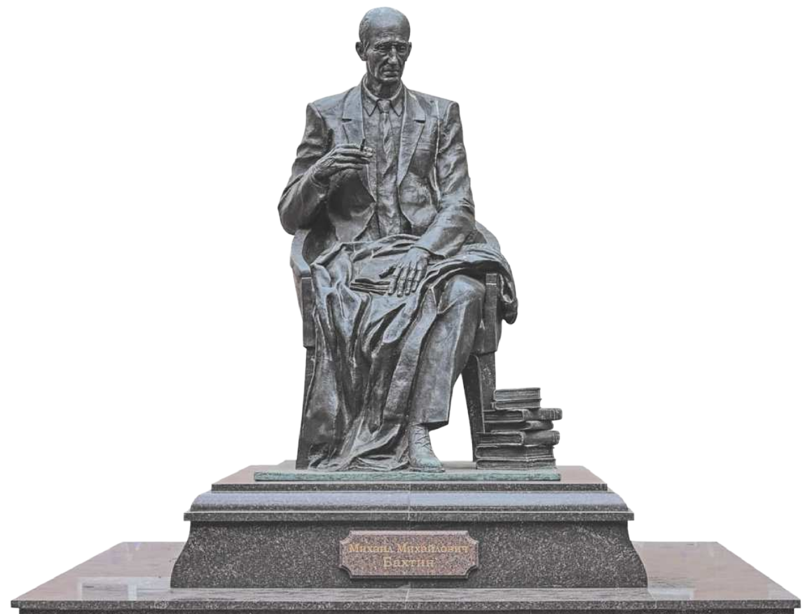 Fotografia da estátua de Mikhail Bakhtin. Ele é um homem careca, de óculos e está sentado em um banco, fumando. Ele usa terno e gravata e sobre seu colo há uma manta e um livro. Ao lado do banco, no chão, há uma pilha de livros.