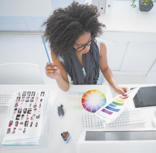 Fotografia de uma mulher sentada a uma mesa de escritório, segurando uma folha com gráficos de cores intensas. Ela observa a folha com uma expressão pensativa.