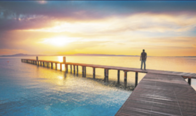 Fotografia de um homem em cima de um convés que avança sobre o mar com o sol se pondo ao fundo. O mar tem a cor vibrante e azul e o céu tons de amarelo intenso.