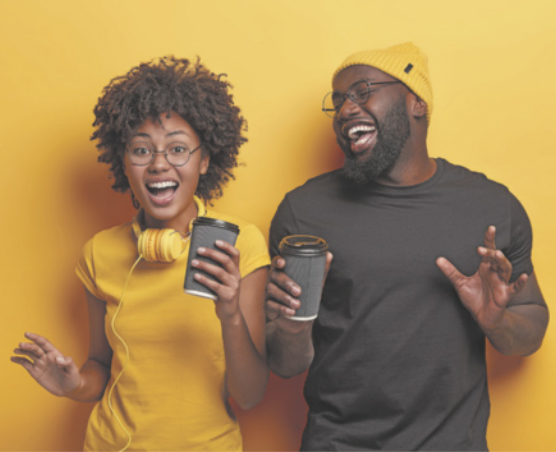 Imagem de dois jovens, um ao lado do outro, em pé, com uma parede amarela ao fundo. Do lado esquerdo, temos uma menina negra de cabelos curtos e encaracolados, usando uma blusa amarela e um fone de ouvido amarelo no pescoço. Ela está com olhos e boca abertos, sorrindo, segurando um copo térmico na altura do rosto. Sua mão direita está abaixada e ela olha para a frente. Ao seu lado, há um homem negro, usando uma touca amarela e uma blusa preta. Ele está com os olhos fechados e a boca aberta, segurando um copo térmico com a mão direita.