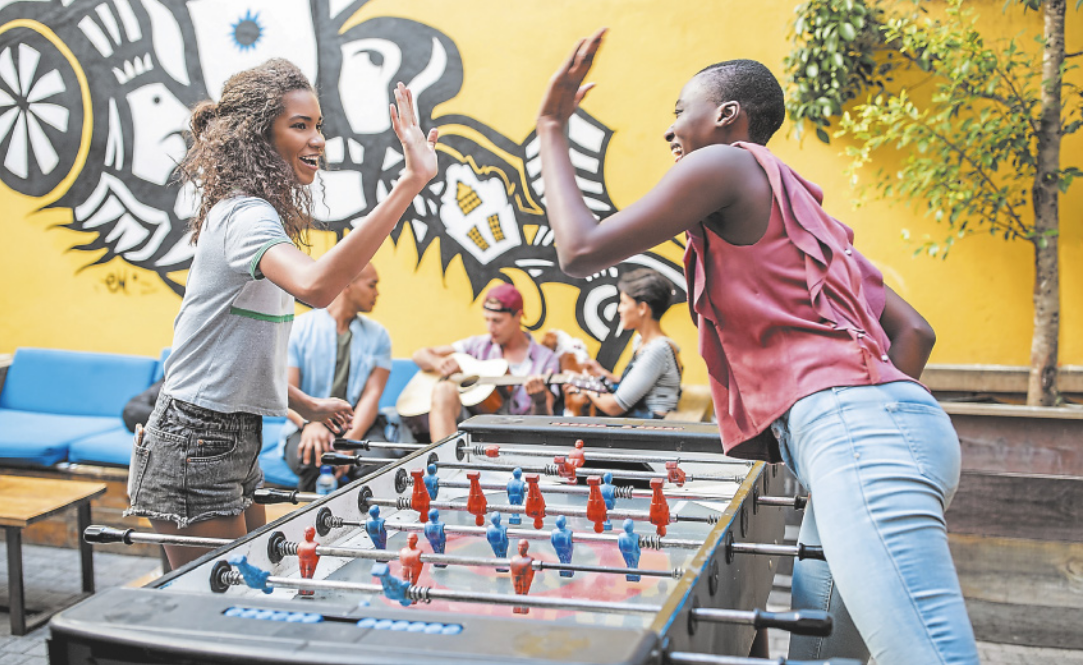 Fotografia de duas jovens em um pátio aberto, jogando pebolim. Elas são negras, uma delas tem o cabelo longo e encaracolado e a outra tem o cabelo curto. Elas estão se cumprimentando com as mãos e, ao fundo, um grupo de jovens toca violão.