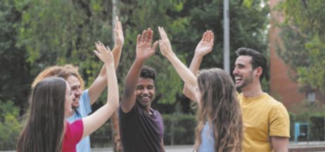 Fotografia de cinco jovens, homens e mulheres, reunidos em um parque. Eles estão em pé, em círculo e erguem uma das mãos.