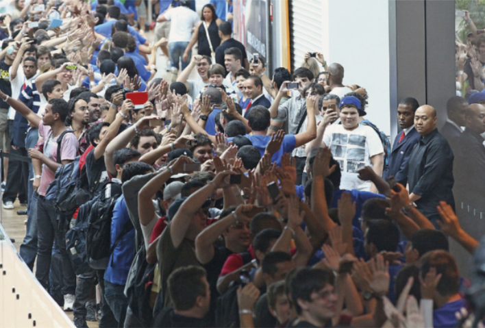 Fotografia de um grande grupo de pessoas formando uma fila longa e compacta. Muitas delas estão com os braços erguidos, acenando ou tirando fotos com seus celulares. Seguranças estão posicionados ao lado da fila, acompanhando o movimento.
