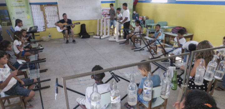 Fotografia de uma sala de aula com crianças sentadas em círculo, participando de uma atividade musical. No centro do círculo, um adulto toca violão, enquanto as crianças o acompanham utilizando instrumentos confeccionados com materiais reaproveitados, como garrafas plásticas e canos.