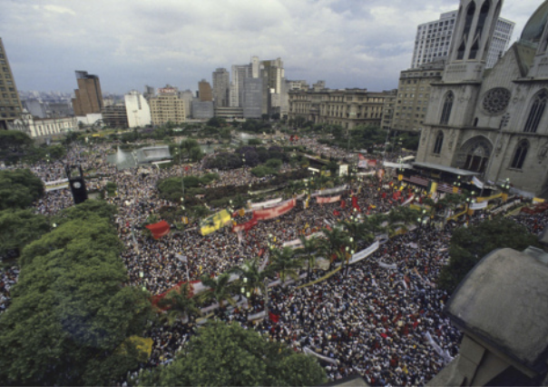 Fotografia aérea de manifestantes ocupando uma grande praça pública em frente a uma catedral.