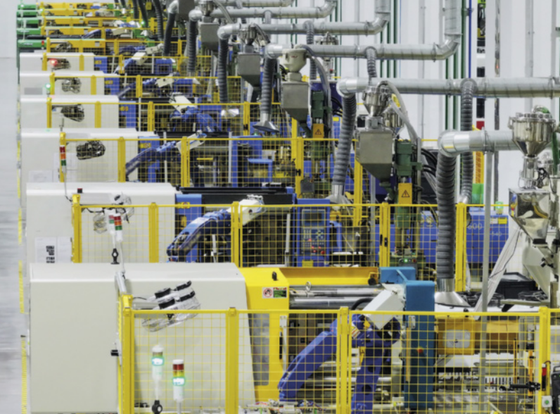 Fotografia do interior de uma linha de produção industrial com várias máquinas organizadas em fileiras paralelas. Cada estação de trabalho está cercada por grades de proteção, e há braços robóticos operando dentro dessas áreas.
