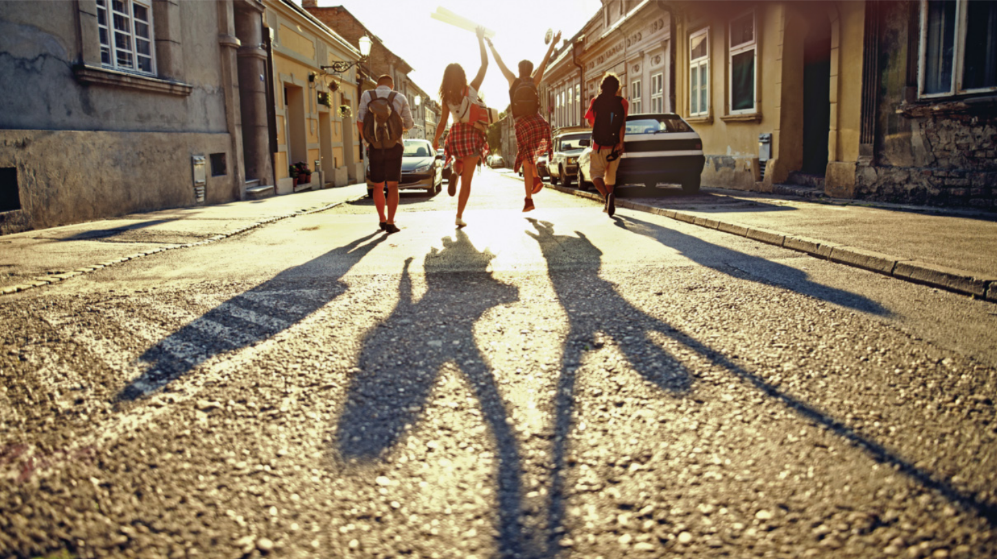Fotografia de quatro jovens, vistos de costas, caminhando por uma rua estreita entre casas antigas. Dois deles, posicionados no centro, pulam com os braços erguidos, enquanto os outros dois caminham pelas laterais. As sombras alongadas de seus corpos se projetam no asfalto.