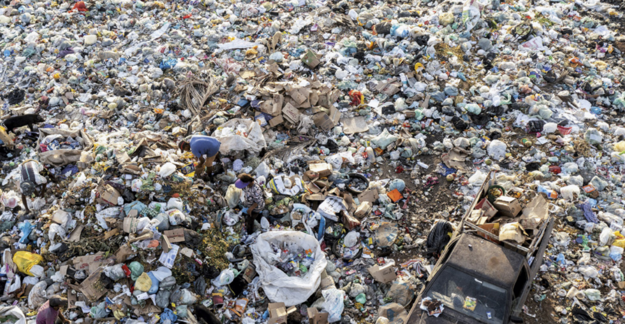 Fotografia de um lixão a céu aberto, com grande quantidade de resíduos espalhados pelo chão, como sacos plásticos, caixas, restos de alimentos e outros materiais descartados. Duas pessoas estão no local, coletando objetos em meio ao lixo. Próximo a elas, há grandes sacos utilizados para armazenar materiais recicláveis.