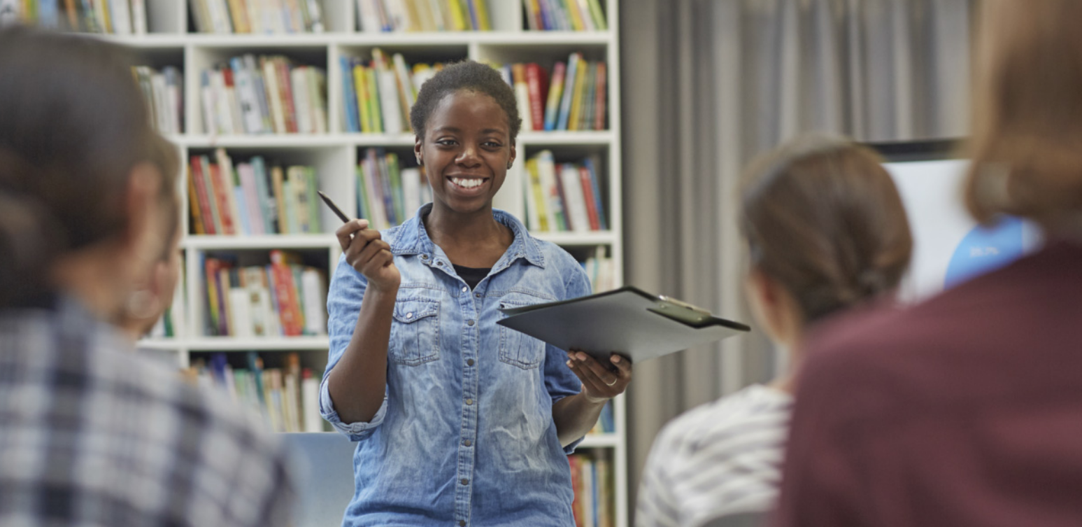 Fotografia de uma mulher negra, jovem, com o cabelo preso, sorrindo enquanto fala para um pequeno grupo de pessoas sentadas, em uma sala onde há uma estante repleta de livros ao fundo. Ela está em pé, vestindo uma camisa jeans e segurando uma pasta preta e uma caneta.