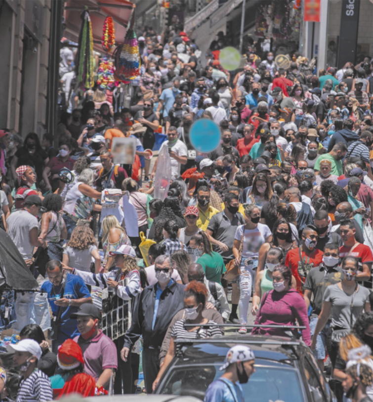 Fotografia de uma rua de comércio popular, extremamente movimentada, com uma grande quantidade de pedestres caminhando em diferentes direções. Muitas pessoas usam máscaras de proteção facial, enquanto outras estão sem. Algumas pessoas carregam sacolas e mercadorias.