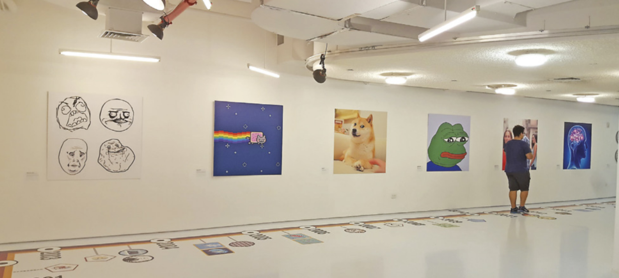 Fotografia de uma galeria de arte contemporânea, onde estão expostas obras dispostas horizontalmente ao longo das paredes. As obras são reproduções de memes populares da internet, incluindo rostos com expressões exageradas, um gato voando e deixando um rastro, um cachorro com expressão curiosa, um sapo com semblante melancólico e um cérebro envolto por luzes. No chão, há uma demarcação que se assemelha a uma linha do tempo. Um visitante observa atentamente as obras expostas.