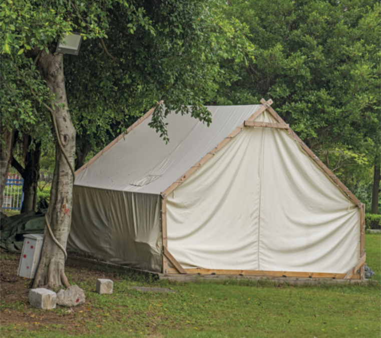 Fotografia de uma tenda montada sobre a grama, em uma área aberta cercada por árvores. A estrutura é simples, composta por vigas de madeira e cobertura de tecido.