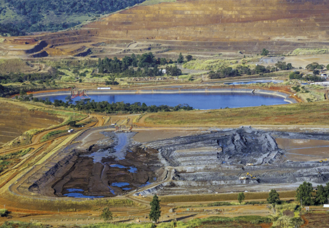 Fotografia de uma área de mineração com escavações em forma de terraços, localizada ao lado de uma represa. O terreno ao redor é aberto, com poucas árvores e uma formação montanhosa ao fundo.