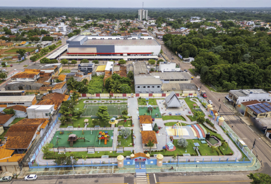 Fotografia aérea de uma cidade, com destaque para uma área de lazer cercada por grades. O espaço conta com diversos equipamentos, como playgrounds, quadras esportivas, áreas cobertas, pistas e caminhos pavimentados. Ao redor, observam-se ruas com veículos, construções residenciais e comerciais, além de uma grande edificação industrial ao fundo e uma extensa área de vegetação à direita.