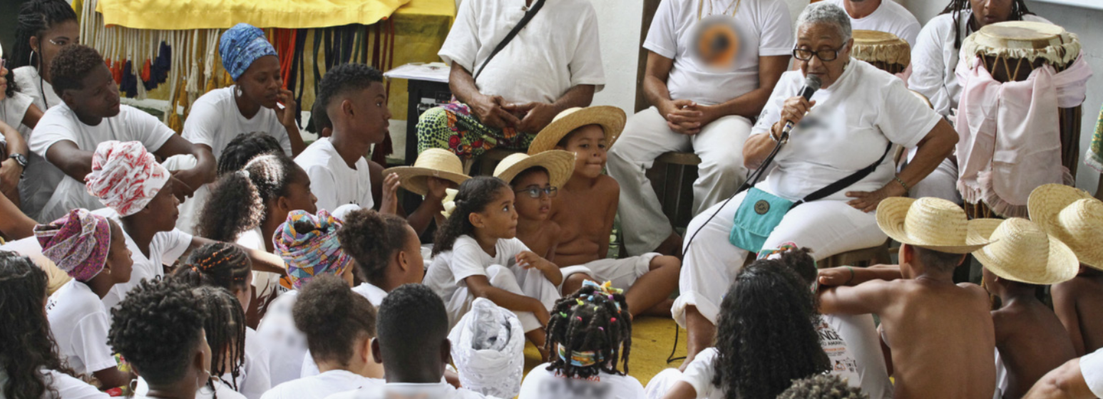 Fotografia de uma mulher negra, idosa, vestida de branco, sentada e falando ao microfone em um altar onde há atabaques e outras pessoas também vestidas de branco, sentadas em cadeiras. Ao seu redor, diversas pessoas estão sentadas no chão, todas vestindo roupas brancas, algumas com turbantes na cabeça e outras com chapéus de palha.
