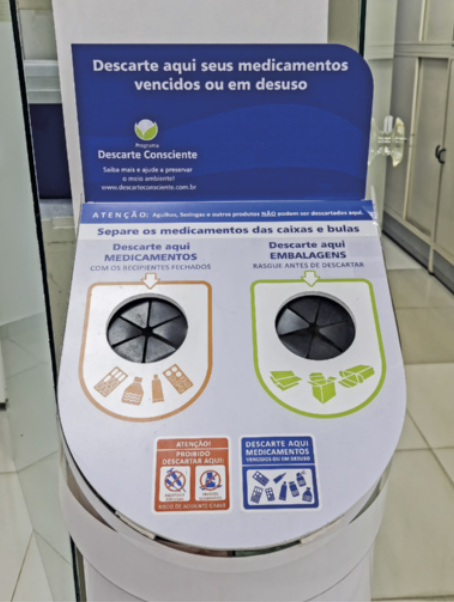 Fotografia de um coletor de medicamentos vencidos. O coletor possui duas aberturas circulares com tampas de borracha flexível, cada uma identificada por orientações sobre o tipo de descarte permitido. Uma abertura é destinada a cartelas, comprimidos e cápsulas; a outra, a frascos, bombinhas e pomadas. Acima das aberturas, há um painel com instruções escritas sobre o descarte correto, informando também quais itens não devem ser depositados no local, como seringas, agulhas e materiais cortantes.