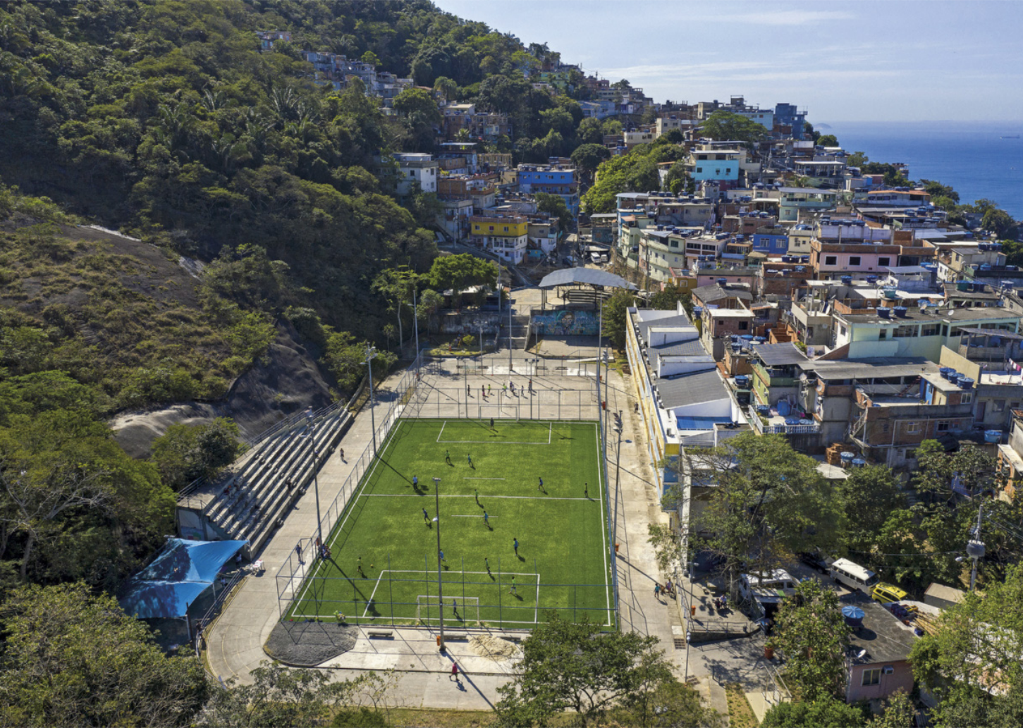 Fotografia aérea de um campo de futebol situado em uma comunidade localizada em um morro. O campo está posicionado na encosta, que é íngreme e coberta por vegetação.