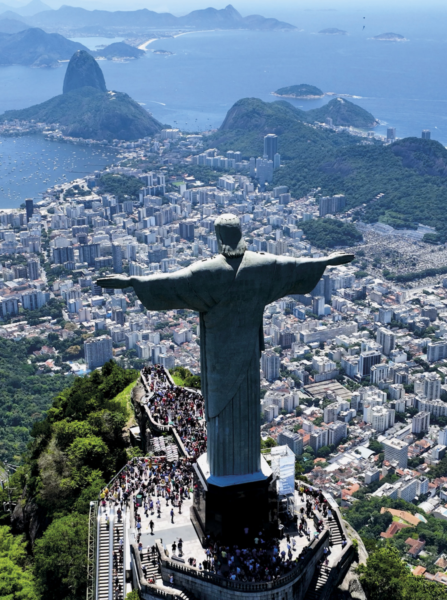 Fotografia aérea de uma grande cidade cercada por morros e pelo mar, com destaque para uma imponente estátua de Jesus Cristo de braços abertos, voltada para a paisagem urbana. A estátua está situada no topo de uma montanha, sobre uma plataforma cercada por numerosos visitantes. Ao fundo, observa-se uma ampla área urbana, com prédios, ruas e áreas de vegetação, além de montanhas e uma baía que abriga diversas ilhas e embarcações.