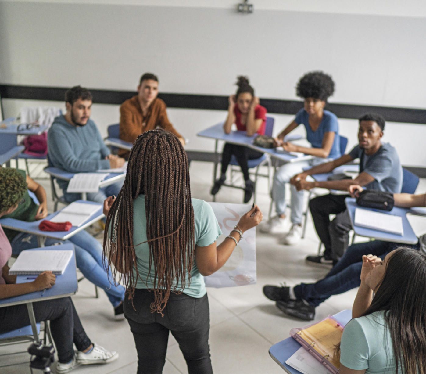 Fotografia de jovens em sala de aula. Uma garota está em pé, mostrando um cartaz para os colegas sentados em carteiras ao seu redor.