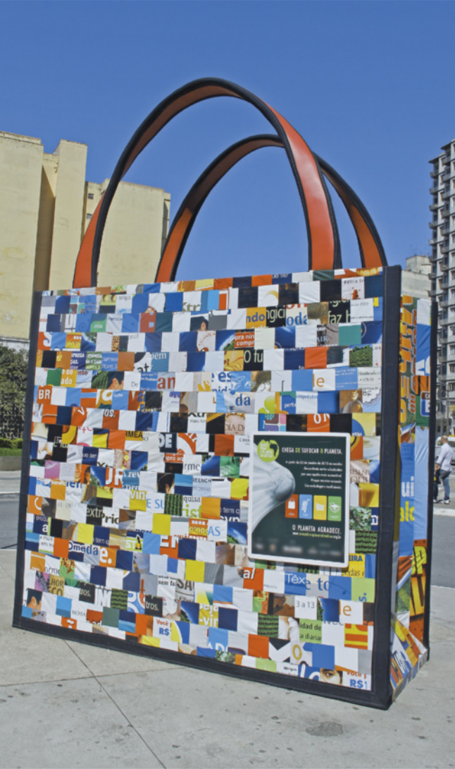 Fotografia de uma grande instalação artística em formato de bolsa de compras retornável, localizada em um espaço público urbano. A superfície da bolsa é composta por recortes de embalagens e rótulos reutilizados, formando um mosaico visual. As alças são feitas de material rígido e curvado. Ao fundo, observam-se prédios e outras estruturas típicas do ambiente urbano.
