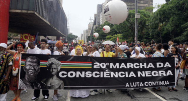 Fotografia de manifestantes caminhando por uma grande avenida, segurando uma faixa com os dizeres: 'Vigésima Marcha da Consciência Negra. A luta continua'.