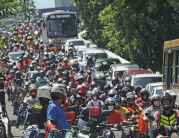 Fotografia de uma grande quantidade de motociclistas ocupando múltiplas faixas de uma movimentada avenida. Muitos deles carregam mochilas ou caixas de entrega nas costas. Os demais veículos ficam restritos a apenas uma faixa da avenida, formando uma grande fila de veículos.