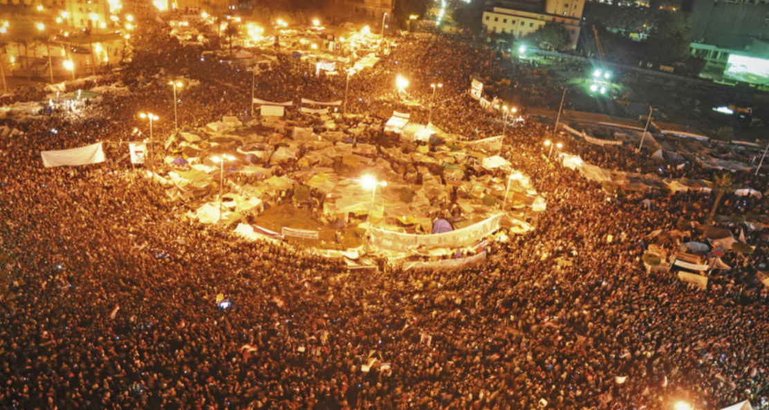 Fotografia aérea de uma grande concentração de pessoas em uma praça, ocupando quase todo o espaço visível. No centro, há tendas e estruturas improvisadas, cercadas pela multidão.