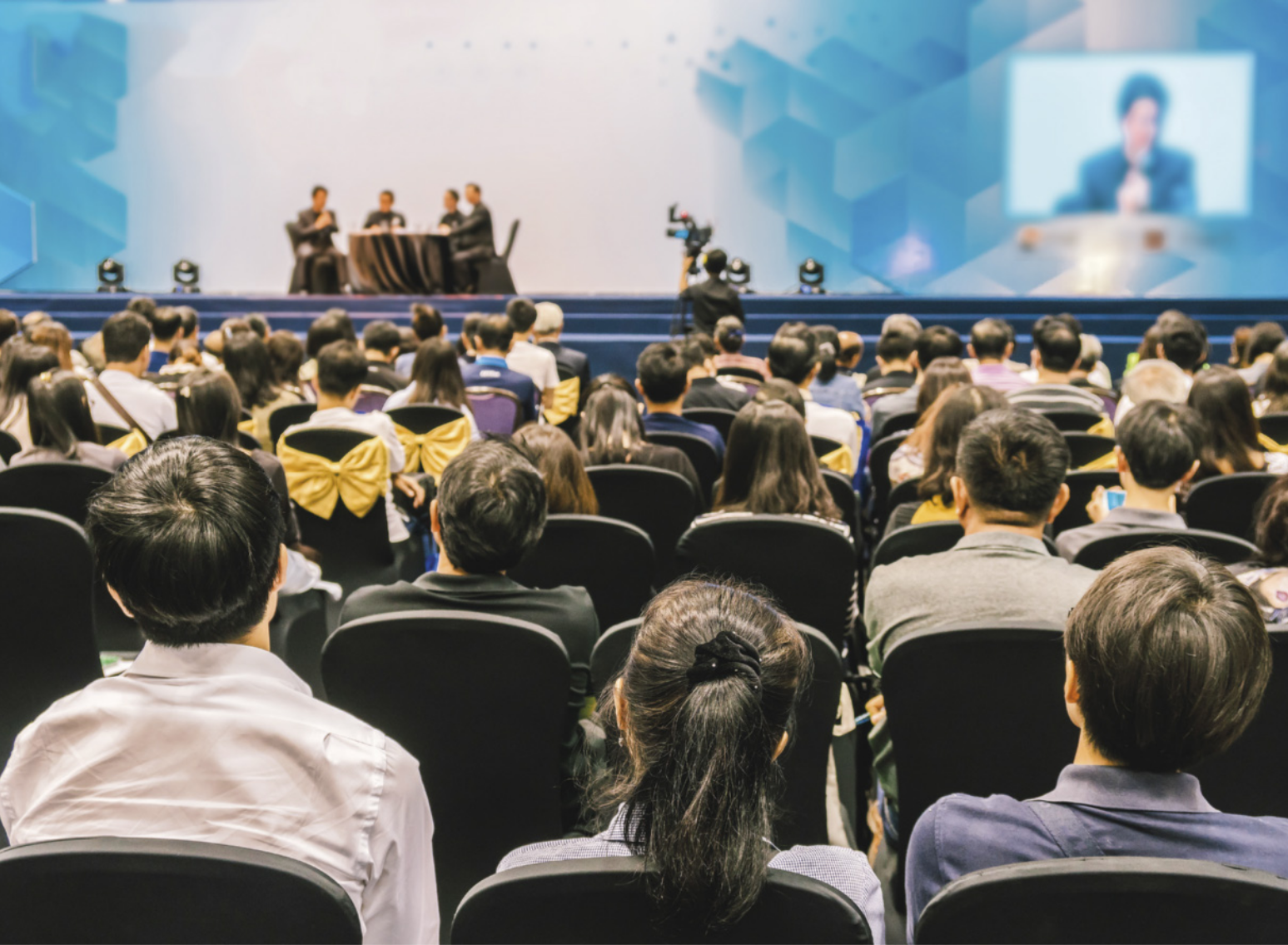 Fotografia de um auditório lotado, com várias pessoas sentadas, assistindo a um evento no palco. Sobre o palco, quatro pessoas estão sentadas em torno de uma mesa redonda. Ao fundo, há um telão que projeta a imagem de uma pessoa.