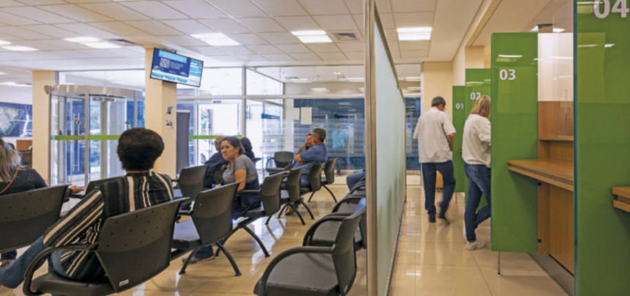 Fotografia do interior de uma agência bancária. À esquerda, várias pessoas estão sentadas em cadeiras dispostas em fileiras. À direita, há divisórias que separam os guichês de atendimento, numerados, onde algumas pessoas estão sendo atendidas.