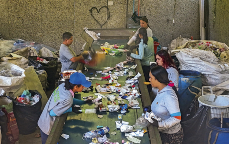 Fotografia de trabalhadores utilizando luvas de proteção enquanto realizam a separação de materiais recicláveis sobre uma bancada. Ao redor, há grandes sacos destinados ao armazenamento dos materiais recicláveis.