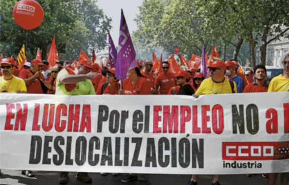 Fotografia de manifestantes caminhando por uma ampla avenida arborizada, carregando bandeiras e segurando, à frente, uma faixa com os dizeres:  'En lucha por el empleo no a deslocalización'.