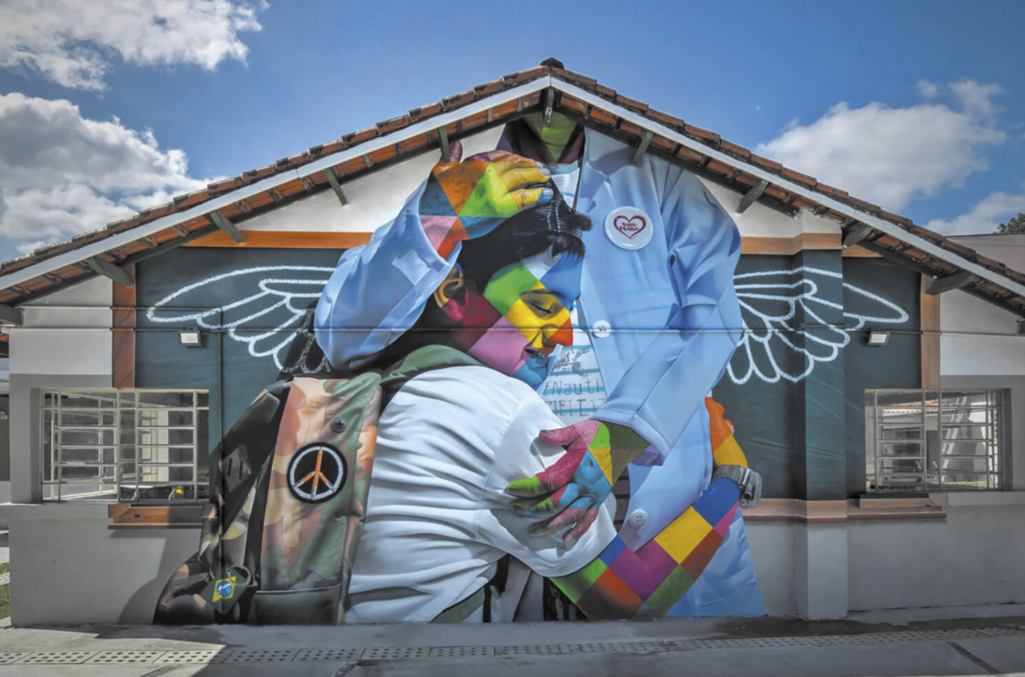 Fotografia da fachada de uma casa com um grande mural pintado. A pintura retrata um estudante com mochila nas costas abraçando uma profissional da saúde, que veste jaleco e máscara. Ao fundo, atrás da figura da médica, há asas de anjo desenhadas em uma lousa escolar.