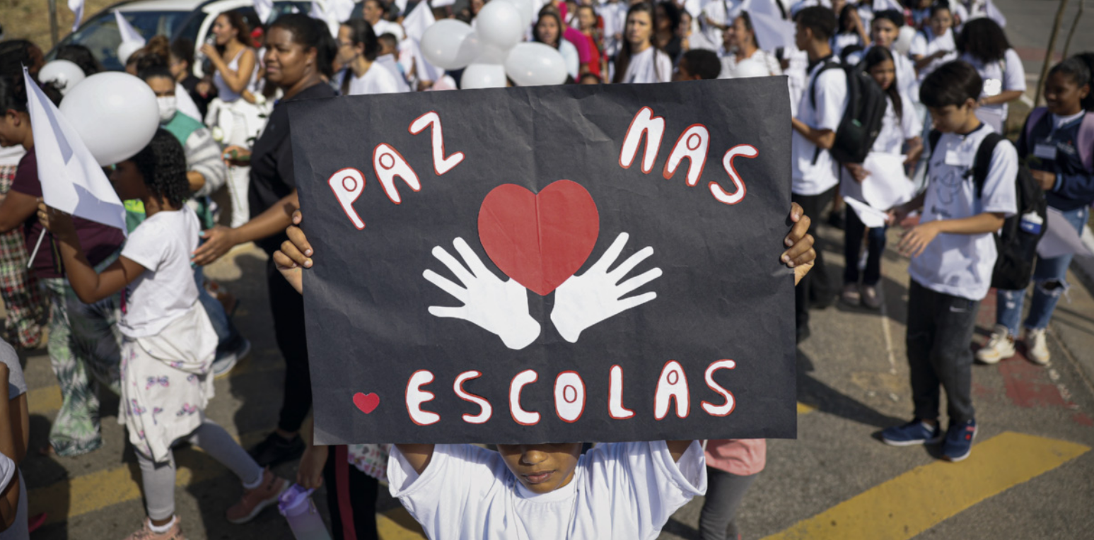 Fotografia de crianças e adolescentes usando camisetas brancas durante uma manifestação em via pública. Uma das crianças segura um cartaz com os dizeres 'Paz nas Escolas' e a imagem de duas mãos lado a lado com um coração no centro.