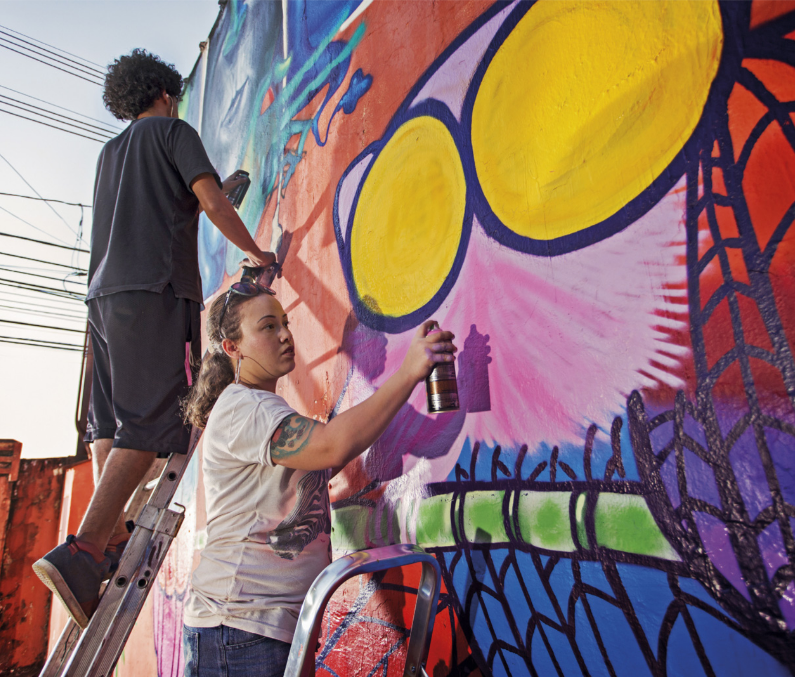 Fotografia de um homem e uma mulher em cima de escadas, grafitando um muro com spray de tinta.