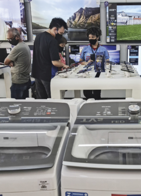 Fotografia do interior de uma loja que vende máquinas de lavar roupa, televisores e celulares. As pessoas presentes utilizam máscaras de proteção e observam os celulares expostos sobre uma bancada.