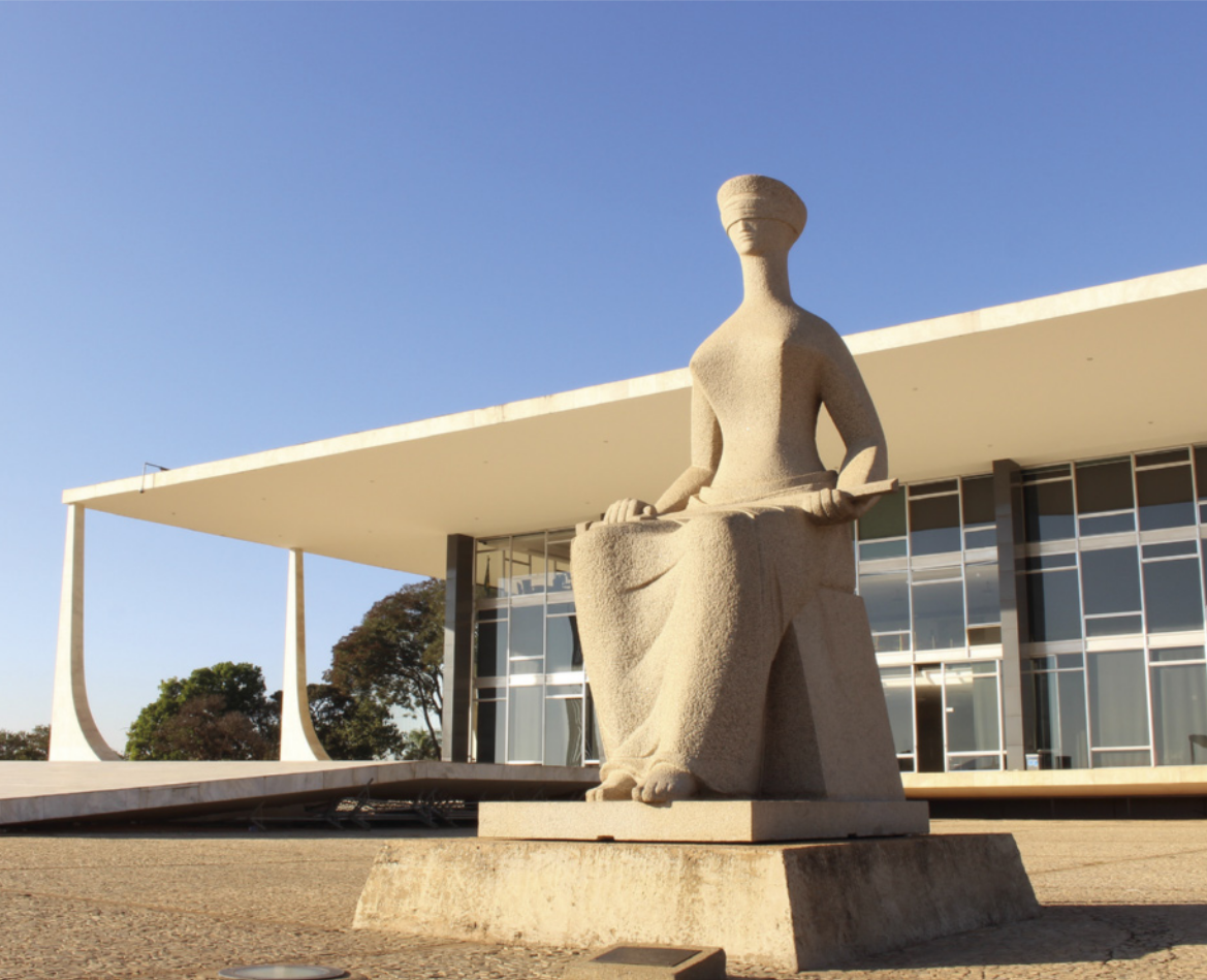 Fotografia de uma estátua de uma figura humana sentada, com os olhos vendados, segurando uma espada sobre o colo. A escultura está posicionada em frente a um edifício de arquitetura moderna, com grandes janelas de vidro e estrutura de linhas retas.
