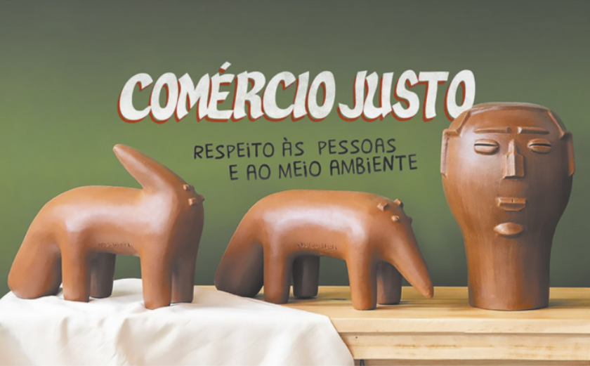 Imagem com três esculturas de cerâmica expostas sobre uma superfície de madeira. Duas das esculturas têm formas que remetem a animais de quatro patas com design abstrato, enquanto a terceira representa uma cabeça humana com traços simplificados. Acima das esculturas, está escrita em destaque a frase: 'Comércio Justo. Respeito às pessoas e ao meio ambiente'.