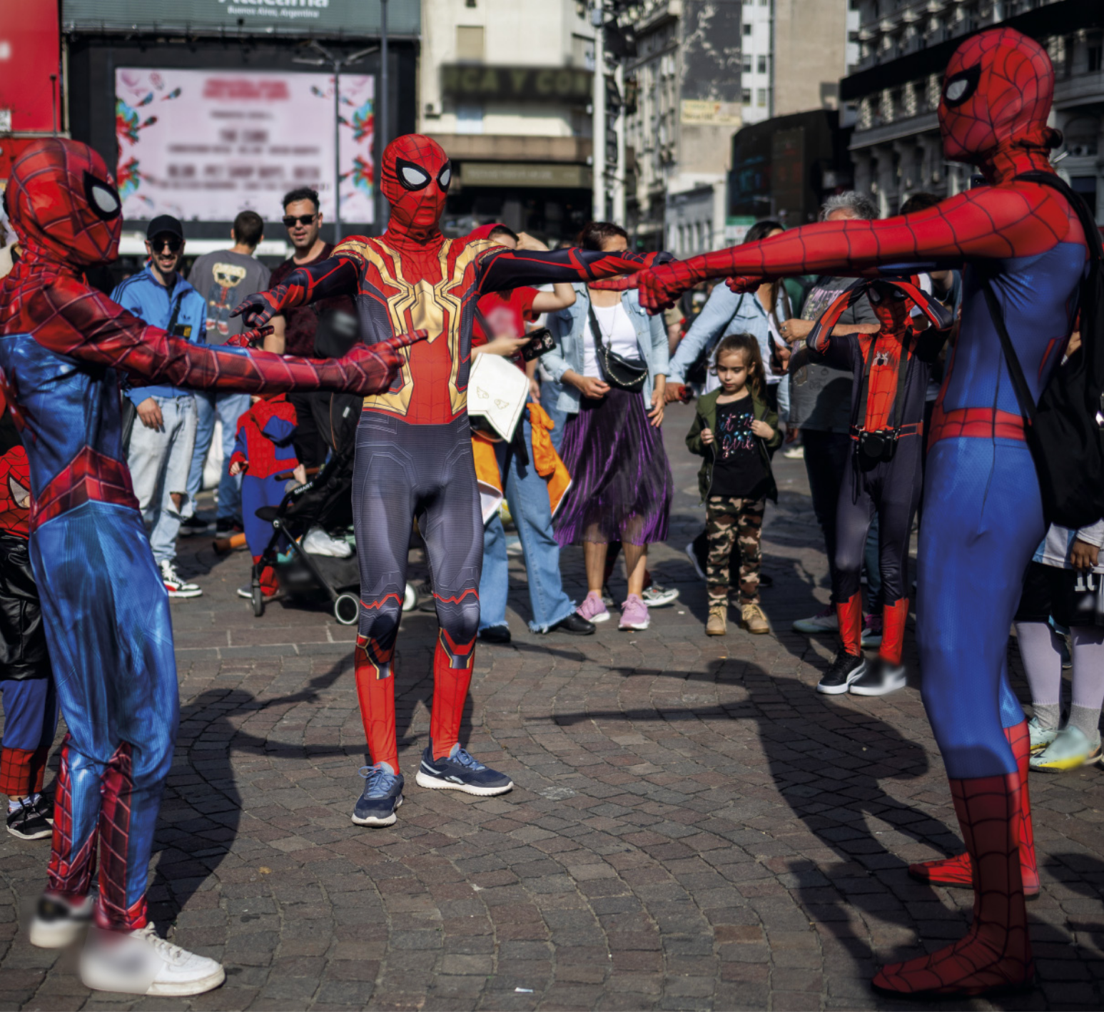 Fotografia de três pessoas fantasiadas de Homem-Aranha, formando uma roda na calçada de uma avenida e apontando umas para as outras, enquanto pessoas ao redor observam a cena.