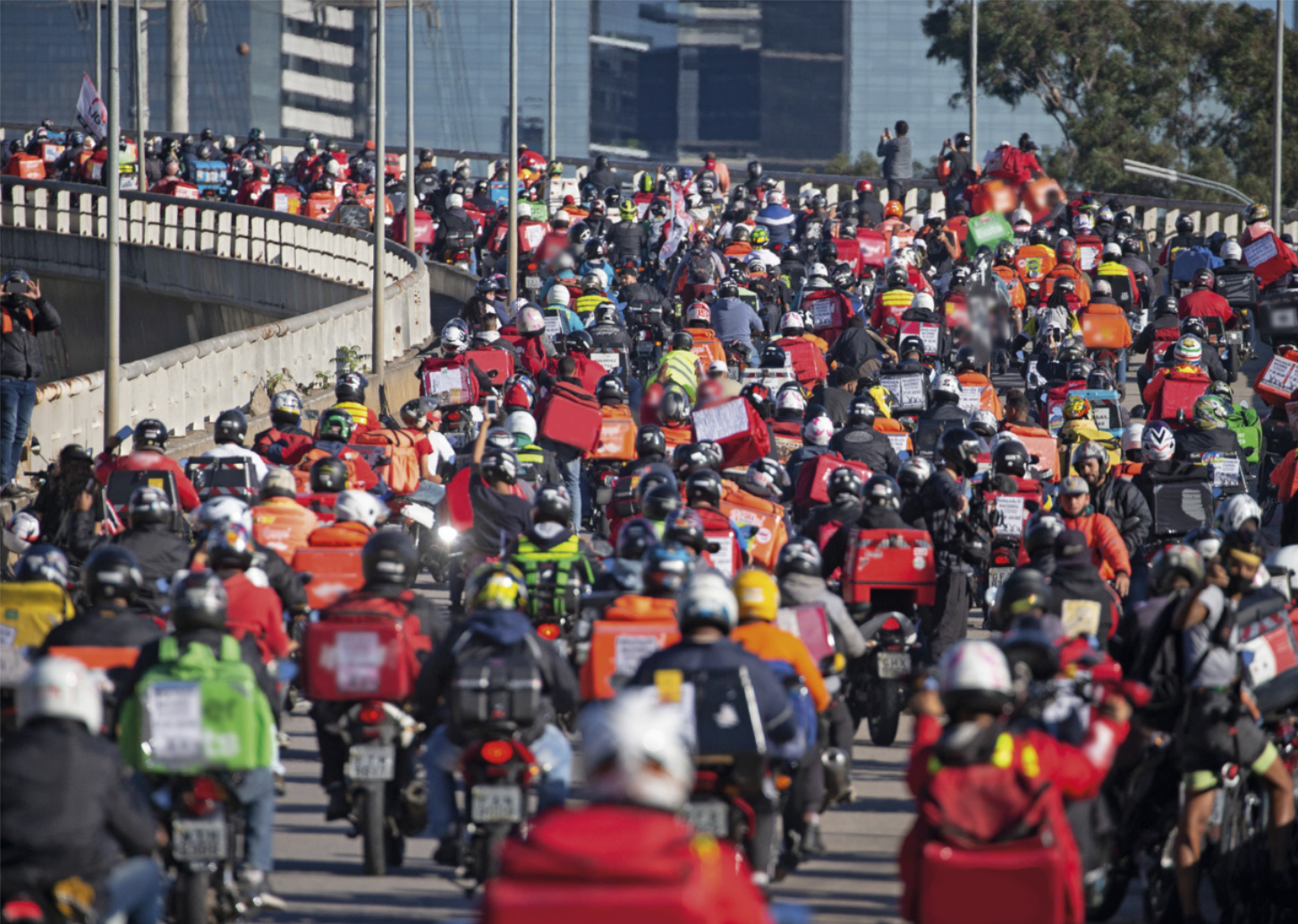 Fotografia de uma grande quantidade de motociclistas ocupando completamente um viaduto em uma área urbana, muitos deles carregando mochilas ou caixas de entrega nas costas.