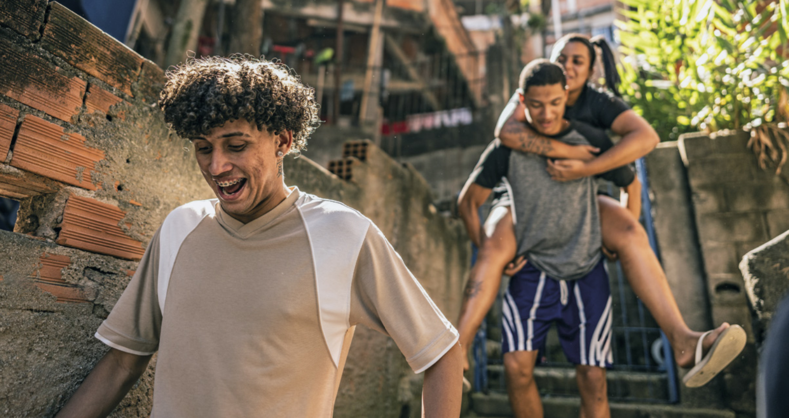 Fotografia de três jovens em um ambiente urbano, com construções simples e escadarias externas. Um rapaz caminha à frente, sorrindo, enquanto, atrás dele, outro jovem desce a escada carregando uma moça nas costas.