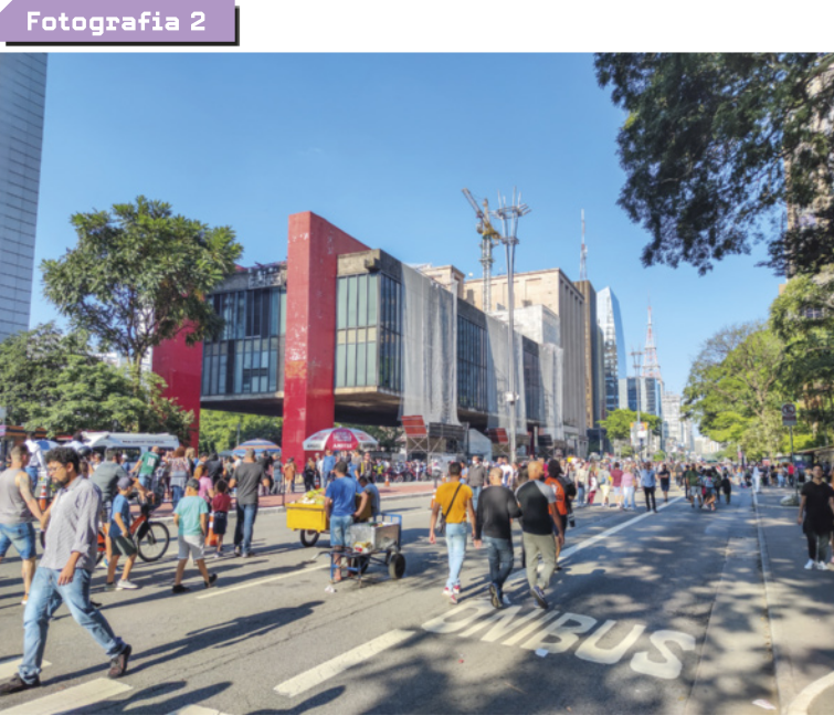 Fotografia dois. Apresenta-se uma avenida por onde as pessoas caminham e andam de bicicleta.