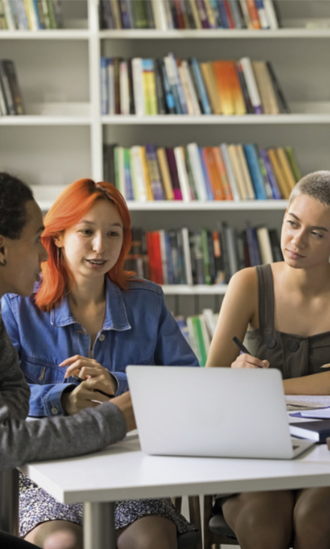Foto de jovens trabalhando juntas. Elas estão sentadas ao redor de uma mesa e fazem anotações usando o computador.