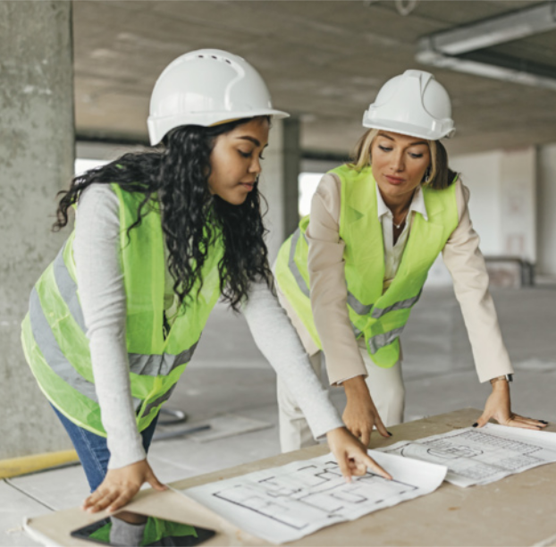 Foto de mulheres analisando uma planta de construção. Elas usam capacetes e coletes de segurança.
