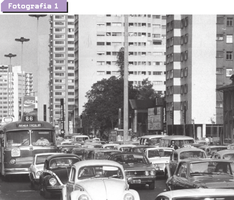 Fotografia um. Apresenta-se uma avenida por onde circulam muitos carros e ônibus.