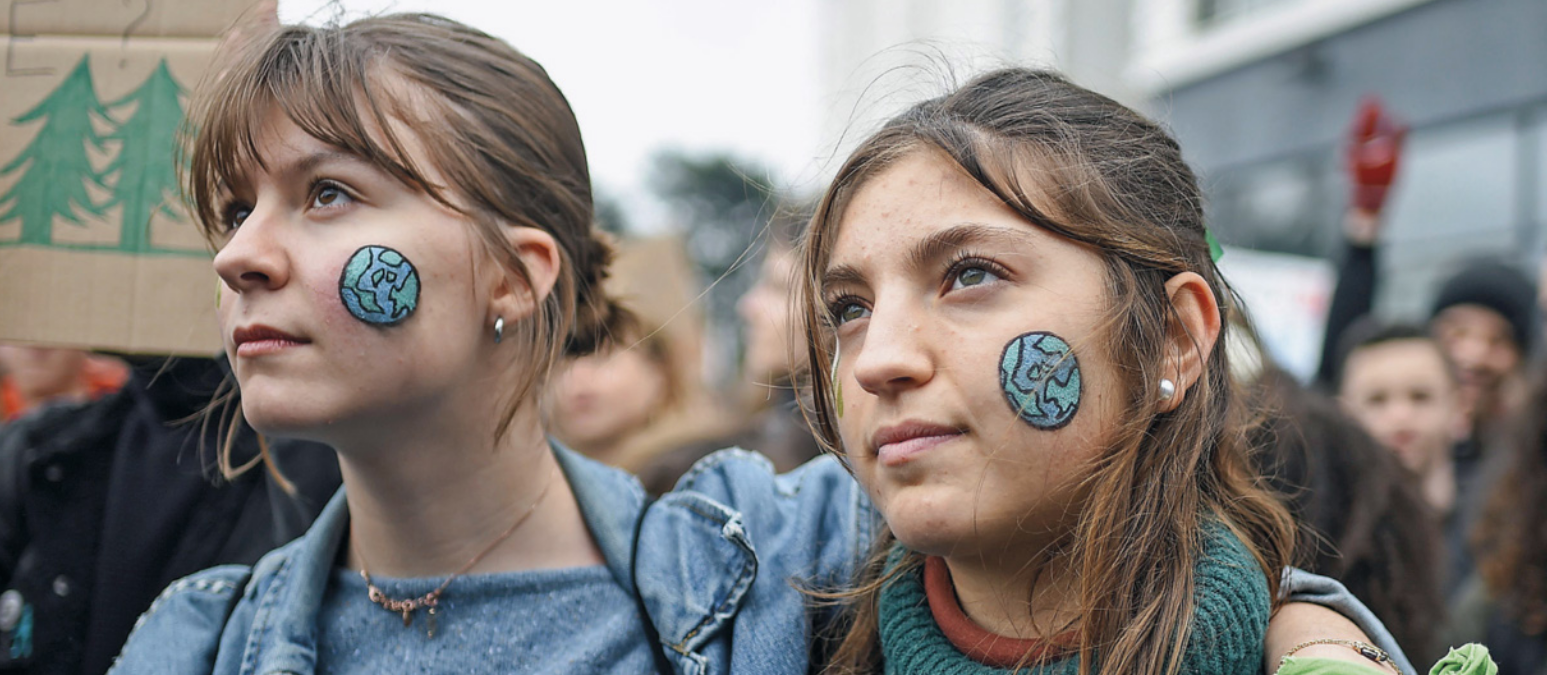 Foto de pessoas em manifestação. Dentre elas, há duas jovens com um planeta Terra pintado no rosto.