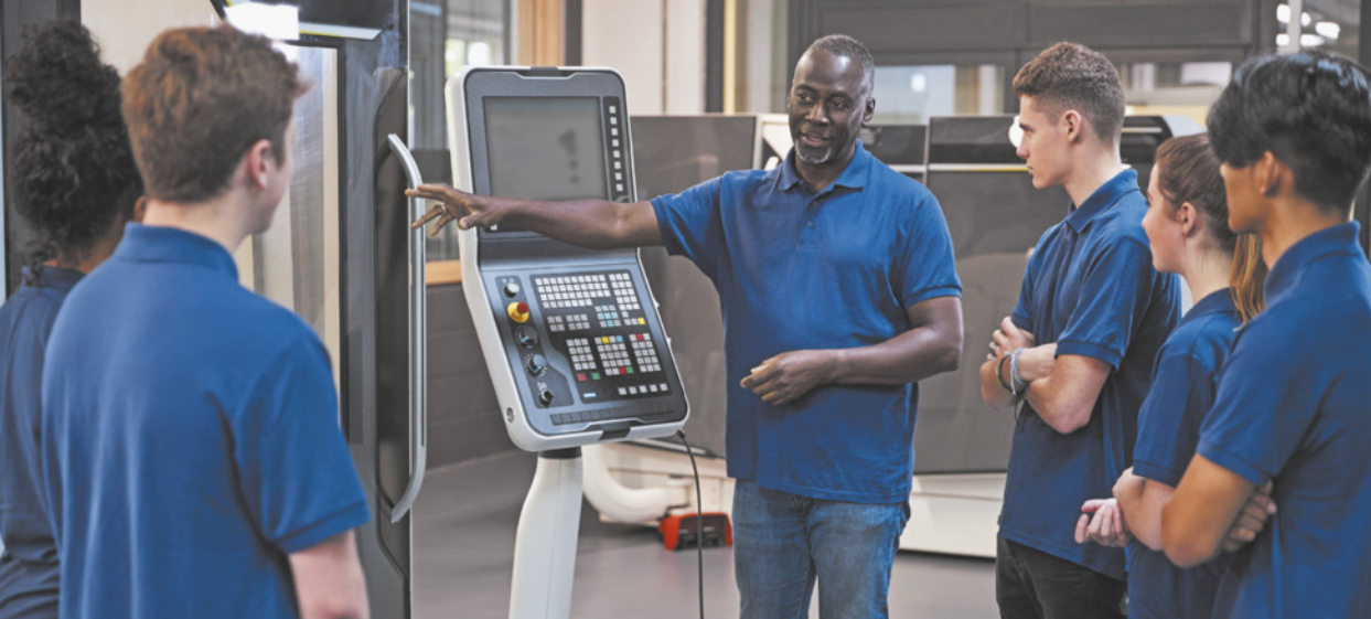 Foto de homem dando instruções para jovens. Ele está em frente a uma máquina enquanto fala.