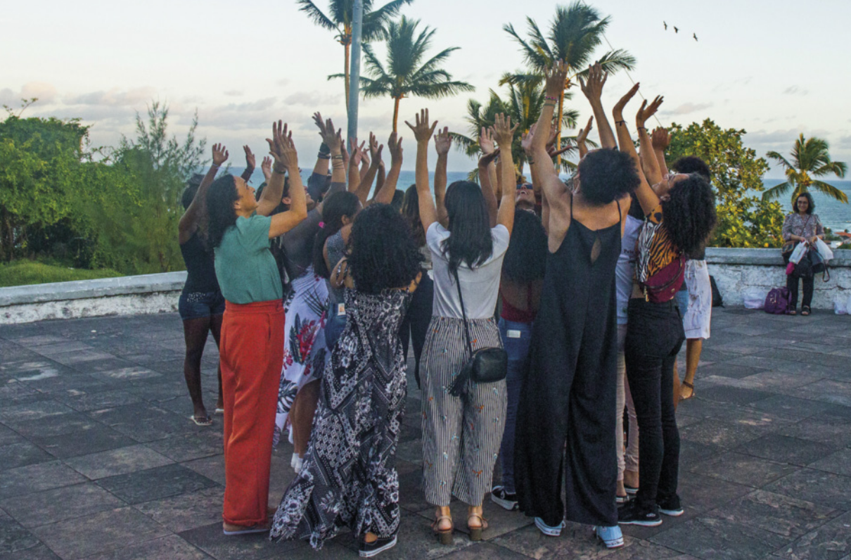 Foto de mulheres em círculo, uma de frente para a outra. Elas erguem os braços em comemoração.