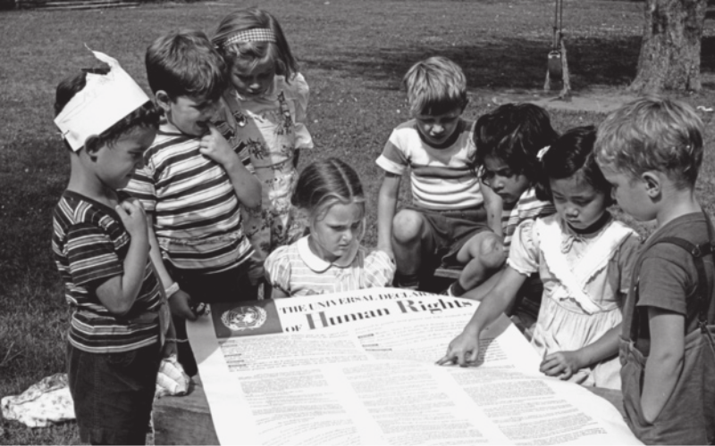 Foto de crianças reunidas ao redor de uma grande folha de papel. Nesta folha está escrita a Declaração Universal dos Direitos Humanos, em inglês.