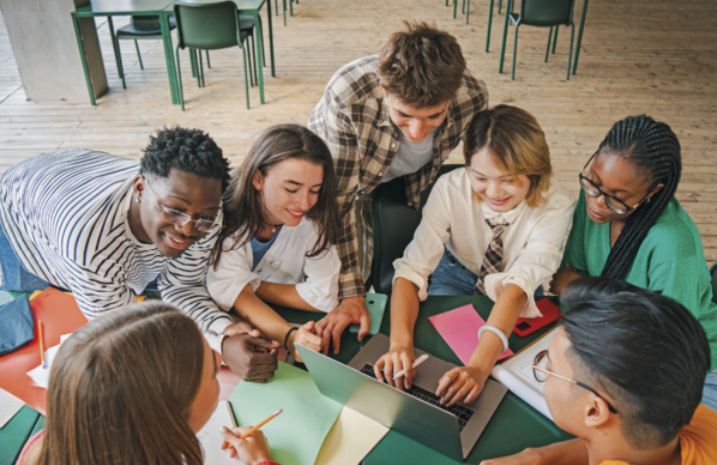 Foto de alunos trabalhando juntos. Eles fazem anotações no caderno e no computador.